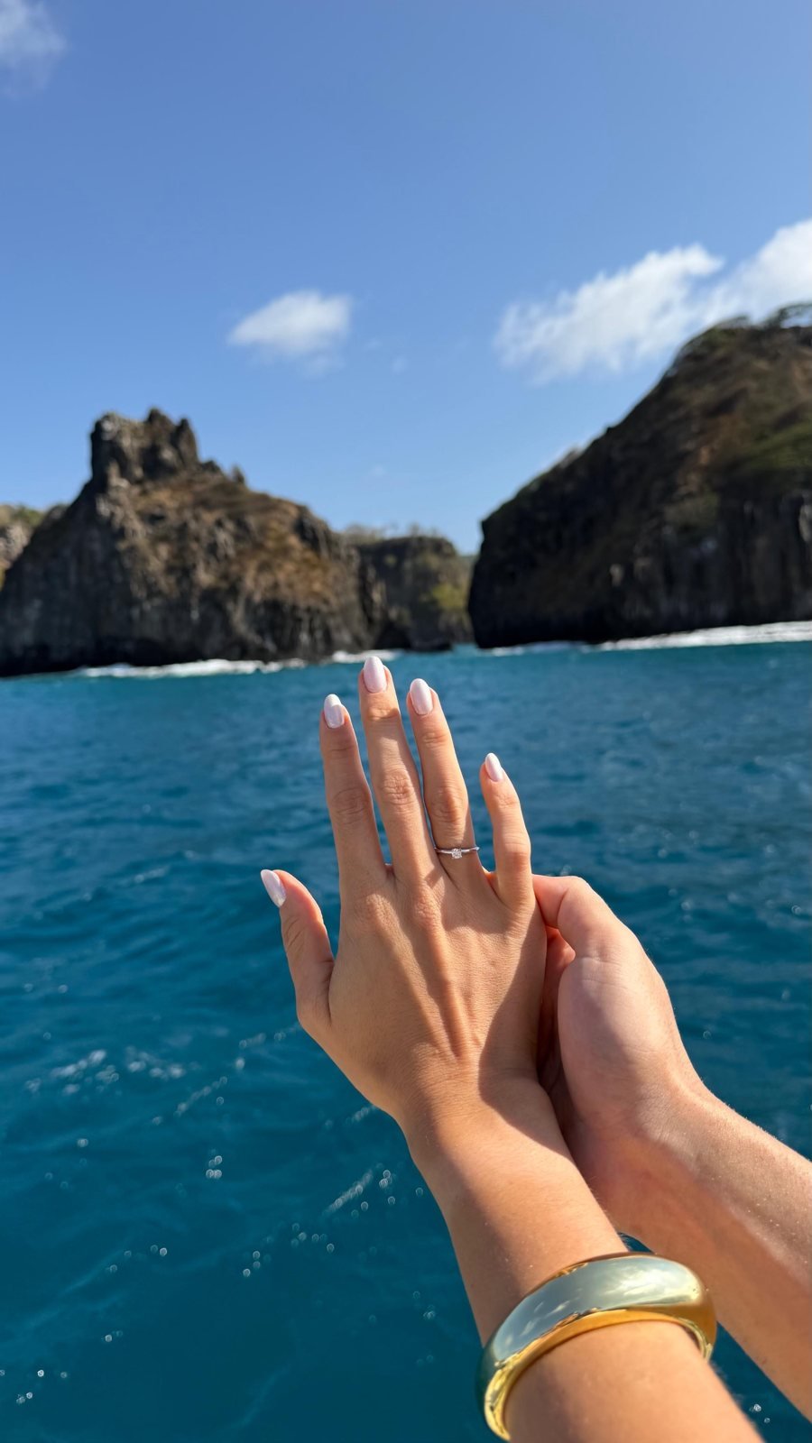 Mão com anel solitário em frente ao mar azul e às rochas de uma ilha