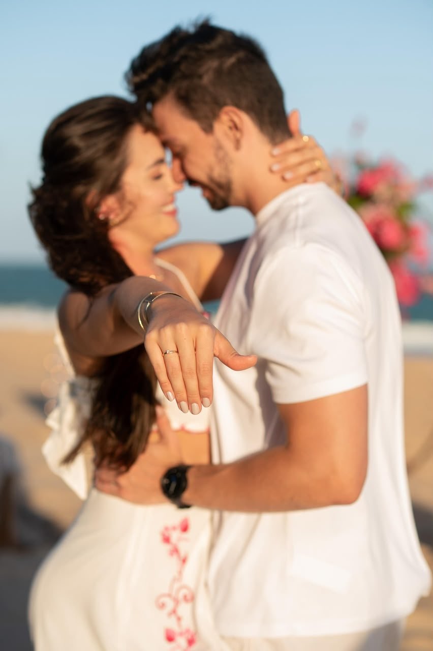 Casal abraçado na praia, com a noiva mostrando o anel de noivado